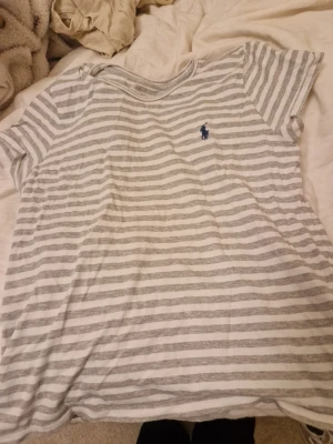 Randig t-shirt från Polo Ralph Lauren - Säljer en vit och ljusgrå randig t-shirt från Polo Ralph Lauren. T-shirten har rund halsringning och en liten blå logga broderad på bröstet. Tillverkad i mjukt bomullsmaterial och har en avslappnad passform. Perfekt för en chill och stilren look.