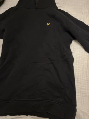Svart hoodie från Lyle & Scott - Svart hoodie från Lyle & Scott med klassisk gul logga på bröstet. Hoodien har en stor känguruficka framtill och en skön huva. Perfekt för en avslappnad och stilren look. Hör av er vid funderingar eller info! Storlek 14-15 years 