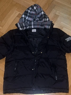 Burberry puffer jacket Large - Snygg svart pufferjacka med Burberry patch på ärmen där det står 'BURBERRY LONDON ENGLAND'. Jackan har dragkedjor framtill, tryckknappar och en huva med grått, vitt och rött rutigt foder. Perfekt för kalla dagar och riktigt trendig. Storlek Large och nyskick. Köpt i andrahand sedan tidigare med tvättlappar avklippta så förmodar att denna inte är autentisk, därav lågt pris.