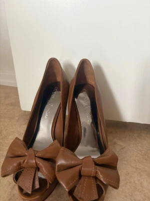 Bruna pumps med rosett från Guess - Snygga bruna pumps från Guess med hög klack och peep-toe. Skorna har en stor dekorativ rosett framtill och är tillverkade i läder. Perfekta för dig som vill sticka ut med en klassisk men trendig look.