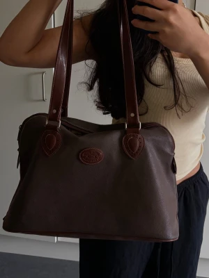 Mörkbrun läderhandväska från The Bridge - Snygg mörkbrun handväska i äkta läder från The Bridge. Väskan har två långa axelband, silverfärgade metalldetaljer och en oval logotyp framtill. Klassisk och rymlig modell med dragkedja upptill och fina sömmar.