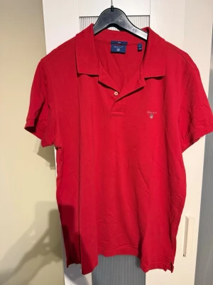 Röd pikétröja från Gant - Klassisk röd pikétröja från Gant med korta ärmar och liten logga broderad på bröstet. Tröjan har krage och två knappar vid halsen. Perfekt till jeans eller shorts för en clean och enkel stil.