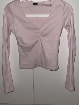 Ljusrosa långärmad topp med v-ringning - Trendig ljusrosa topp med lång ärm och snygg v-ringning. Toppen har en rynkad detalj framtill som ger en cool twist. Perfekt att matcha med jeans eller kjol för en enkel men stilsäker look.