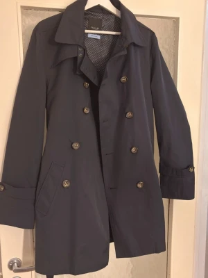 Helt ny Mörkblå trenchcoat från sand Copenhagen  - Stilren mörkblå trenchcoat från sand Copenhagen  med dubbelknäppning, klassisk krage och bälte i midjan med metallspänne. Jackan har sidofickor och dekorativa knappar vid ärmsluten. Perfekt för dig som vill ha en tidlös och snygg look.