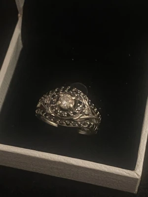 Silverring med pärldetaljer - Unik silverring med en rund, klar sten i mitten och små pärldetaljer runt om. Ringen har ett antikt inspirerat mönster och är tillverkad i silverfärgat metall. Perfekt för dig som gillar detaljerade och klassiska smycken.