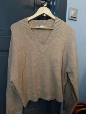 Beige stickad tröja med v-ringning från Zara - Säljer en beige stickad tröja från Zara med djup v-ringning och breda ribbade kanter vid hals och nederkant. Tröjan har en avslappnad passform och långa ärmar, perfekt för att skapa en chill och snygg look. Materialet är mjukt och känns skönt mot huden.
