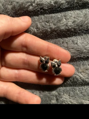 Söta uggleörhängen i silverfärg - Ett par örhängen formade som ugglor i silverfärgad metall med svarta stenar som ögon och mage. De har en glittrig detalj runt magen och är små och nätta, perfekta för att ge en unik touch till din stil.