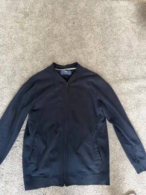 Marinblå tröja Dressmann - Snygg marinblå tröja från Dressmann med dragkedja framtill och två sidofickor. Jackan har ribbade muddar vid hals, ärmslut och nederkant. Tillverkad i mjukt bomullsmaterial för en avslappnad look.