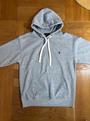 Grå hoodie från Polo Ralph Lauren - Snygg ljusgrå hoodie från Polo Ralph Lauren med vit dragsko i huvan och klassisk känguruficka framtill. Liten mörkblå logga broderad på bröstet. Tillverkad i mjuk bomullsblandning, perfekt för chill dagar eller streetwear-stil. Storlek M men sitter som S