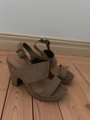 Beigea Klackar med bred klack - Snygga pumps i beige mockaimitation med breda remmar och chunky klack. Justerbart spänne vid ankeln och öppen tå ger en trendig look. Perfekta för dig som gillar höga klackar och vill sticka ut lite extra.💕Ganska använda på fram på trampdynan, men syns ej vid användning.