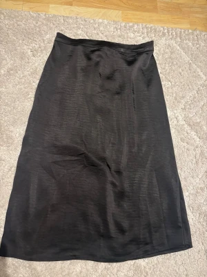 Svart långkjol i satin från Vila - Säljer en svart långkjol från Vila i glansigt satinmaterial. Kjolen har en enkel, rak passform och resår i midjan för extra komfort. Perfekt att styla med både sneakers och boots för en trendig look.