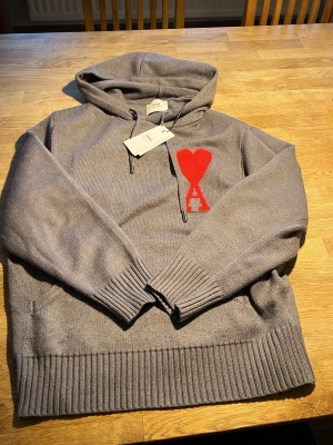 Grå hoodie från Ami Paris med röd logga - Säljer en grå stickad hoodie från Ami Paris med stor röd hjärtlogga och bokstaven A på bröstet. Hoodien har ribbade muddar, ficka framtill och dragsko i huvan. Materialet är mjukt och tjockt, perfekt för kyliga dagar. Snygg och trendig design!