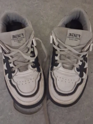 Vita sneakers från Beier - Säljer ett par vita sneakers från Beier med svarta detaljer och beige snören. Skorna har en klassisk sportig design med perforeringar på tån och en tjock sula. Märket syns tydligt på plösen och de har en bekväm passform för vardagsbruk.