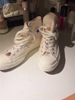 Beige Converse Chuck Taylor High - Säljer ett par klassiska Converse Chuck Taylor All Star high tops i beige canvas med vita gummisulor och tåhättor. Skorna har vita skosnören, silverfärgade öljetter och den ikoniska Converse-loggan på sidan. Perfekta för en avslappnad och trendig stil. Helt nyinköpta och oanvända! 