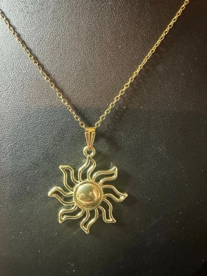 Guldigt halsband med solhänge - Snyggt halsband i guldfärgad metall med ett stort solformat hänge. Hänget har en rund mitt och vågiga solstrålar som ger en cool och unik look. Kedjan är tunn och elegant, perfekt för att lyfta vilken outfit som helst. Märke och material är ej angivet.