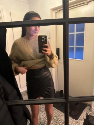 Svart omlottkjol i skinnimitation - Snygg svart kort omlottkjol i skinnimitation med lätt glansig finish. Kjolen har en figurnära passform och omlott-detalj framtill som ger en trendig look. Perfekt att styla med en stickad tröja eller t-shirt.