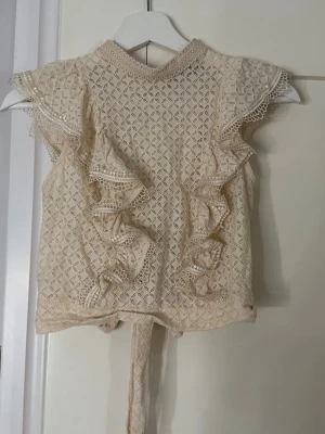 Beige spetsblus med volanger - Supersöt beige topp i spets med volangdetaljer framtill och på ärmarna. Toppen har korta ärmar, knytband i midjan och ett fint hålmönster över hela plagget. Perfekt för dig som gillar romantisk och feminin stil.