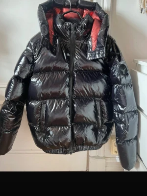 Svart pufferjacka från Moncler - Säljer en svart pufferjacka från Moncler med glansigt material och röd innerfoder. Jackan har hög krage, avtagbar huva och stora fickor framtill. Perfekt för kalla vinterdagar och riktigt snygga att bära. Moncler-logga på ärmen.