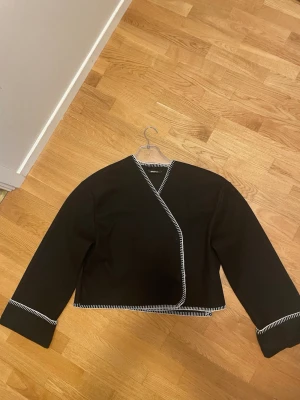 Svart omlottkofta med vita detaljer - Syrorlek L men sitter som S-M. Stilren svart omlottkofta med vita kontrastsömmar längs kanterna och ärmsluten. Koftan har en croppad passform och raka, långa ärmar. Perfekt för att lyfta en enkel outfit med en unik twist. Båda knapparna fungerar ej tyvärr men går att använda som en kofta som man slänger över liksom, ena knappen är helt av 