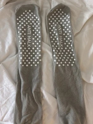 Grå halksockor med anti-halk - Dubbel sidiga Mysiga grå halksockor med vita gummiprickar undertill för extra grepp. Insidan är mjuk och frottéliknande för skön komfort. Perfekta för att glida runt hemma utan att halka. Texten 'ANTI HALK' på undersidan. Snygg och enkel design.