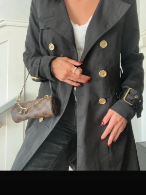 Svart trenchcoat med guldknappar - Stilren svart trenchcoat med stora guldknappar och bred krage. Jackan har bälte med spänne vid ärmsluten och klassisk dubbelknäppning framtill. Perfekt för dig som vill ha en tidlös och trendig look.