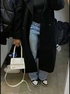 Svart lång trenchcoat med bälte - Stilren svart trenchcoat med klassisk krage och knytskärp i midjan. Lång modell med markerad midja och långa ärmar. Perfekt för en trendig och elegant look. Material 100% polyester 