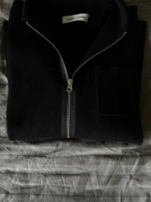 Svart ziptröja från Samsøe Samsøe - Stilren svart ziptröja från Samsøe Samsøe med hel dragkedja framtill och en bröstficka. Tröjan har lång ärm och hög krage, perfekt för lager på lager. Materialet ser ut att vara mjukt och lite tjockare, vilket ger en skön känsla.