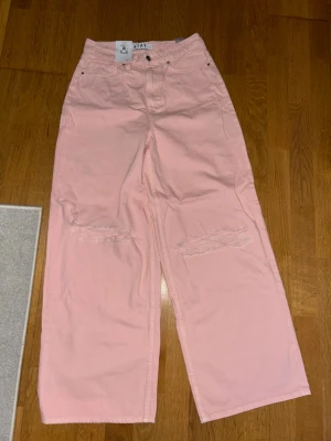 Ljusa rosa wide leg jeans från Stay - Säljer ett par ljusrosa jeans från Stay med breda ben och hög midja. Jeansen har slitna detaljer över knäna och klassisk femficksmodell. Perfekta för dig som vill ha en avslappnad och trendig look. Materialet är bomull. I storlek S.