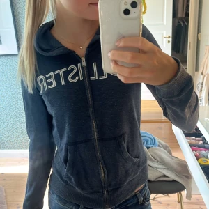 Mörkblå hoodiejacka från Hollister - Snygg mörkblå hoodiejacka från Hollister med vit broderad logga över bröstet. Jackan har dragkedja framtill, och ficka vid magen.