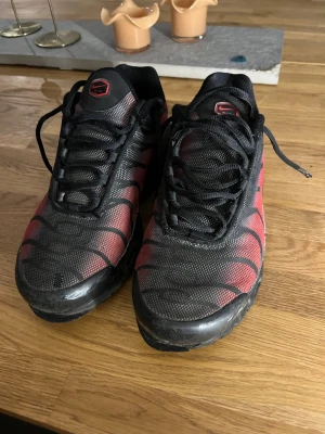 Nike tns - Säljer ett par Nike tns sneakers i svart och rött med klassiska vågmönster på ovandelen. Skorna har svarta snören, meshmaterial och synlig Air-dämpning i sulan. Perfekta för dig som gillar streetwear och vill sticka ut med en ikonisk modell. Lite sönder längst fram 