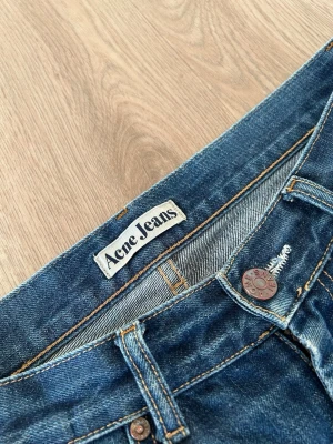 Blå Acne Jeans med raka ben - Säljer ett par klassiska blå jeans från Acne Jeans med raka ben och snygga slitningar. Jeansen har fem fickor, kopparfärgade nitar och kontrastsömmar. Perfekta för en avslappnad och trendig look.