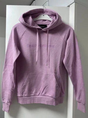 Ljuslila hoodie från Peak Performance - Säljer en ljuslila hoodie från Peak Performance med broderad logga på bröstet. Hoodien har huva med dragsko, magficka och ribbade muddar vid ärmslut och nederkant. Perfekt för en avslappnad och snygg stil.