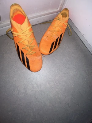 Adidas X Speedportal FG Orange stl 44.5 - Säljer ett par orangea fotbollsskor från Adidas med svarta detaljer och klassiska ränder på sidan. Skorna har snörning och är tillverkade i ett syntetmaterial som ger bra grepp och känsla på planen. Perfekta för dig som vill sticka ut på matchen!Nypris: ungefär 1500–1700 kr.