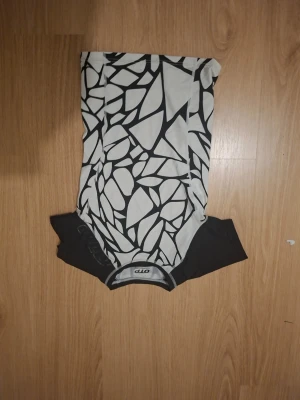 Svartvit topp med grafiskt mönster - Cool topp från Aio med svartvita grafiska mönster som liknar ett nät eller mosaik. T-shirten har korta ärmar och rund halsringning. Perfekt för dig som gillar unika prints och vill sticka ut.