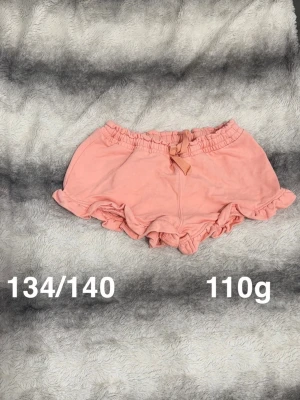 Aprikosfärgade volangshorts med rosett - Söta aprikosfärgade shorts med volangkant och en liten rosett framtill i midjan. Shortsen har elastisk midja och är perfekta för varma dagar. 🌸 Fint använt skick. Storlek 134/140 