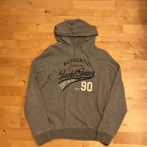 Grå hoodie från Jack & Jones - Snygg grå hoodie från Jack & Jones med tryck på bröstet i svart och vitt. Klassisk modell med huva och ribbade muddar vid ärmslut och nederkant. Perfekt för en avslappnad och stilren look.