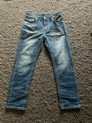 True Religion blå jeans Joey super T modell - Säljer ett par blå jeans från True Religion med tydliga vita kontrastsömmar och snygga detaljer på fickorna. Modellen har klassiska fem fickor, knappar i kopparfärg och logopatch bak. Materialet är jeans och passformen är rak med normal midja. Nästan nyskick då jag använt dom en endaste gång. 