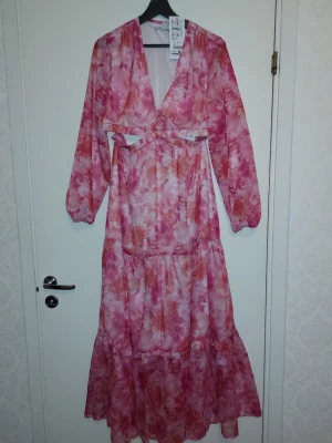 Lång blommig klänning med cut out - Lång klänning i rosa och orange toner med blommigt mönster. Djup v-ringning, långa ärmar och trendig cut out-detalj i midjan. Flödande material som ger en luftig känsla, perfekt för varma dagar.