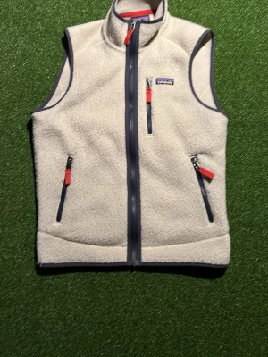 Vit fleeceväst från Patagonia - Säljer en krämvit fleeceväst från Patagonia med hel dragkedja framtill och tre fickor med kontrasterande mörkblå dragkedjor och röda detaljer. Västen har hög krage och mjukt, fluffigt material som ger en avslappnad look.