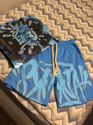 Blå shorts från SikSilk med snörning - Säljer ett par blå shorts från SikSilk med stort graffiti-inspirerat tryck i ljusblått. Shortsen har bred resår i midjan och grov vit snörning framtill. Perfekta för en avslappnad streetwear-look.