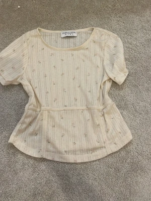 Beige blommig topp Unique Collection - Söt beige topp från Unique Collection med diskret blommigt mönster och tunna vertikala ränder. Toppen har rund halsringning, korta puffärmar och markerad midja för en feminin siluett. Perfekt att styla med jeans eller kjol. storleken står inte men jag tycker själv den passar som 146/152