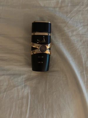 Lattafa Asad parfym svart och guld - Lattafa Asad parfymflaska i matt svart med eleganta gulddetaljer och arabiska bokstäver. Flaskan har en unik, modern design med guldband runt kroppen och ett dekorativt emblem framtill. Perfekt för dig som gillar exklusiva och stilrena dofter.