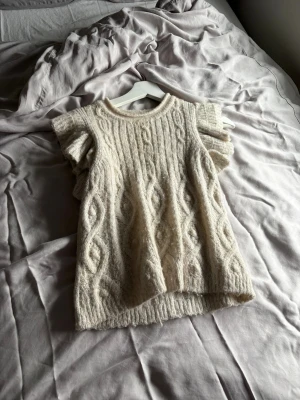 Vit stickad topp från Jessica Simpson - Supersöt vit stickad topp från Jessica Simpson med korta volangärmar och kabelstickat mönster. Mjuk och fluffig känsla, perfekt för lager på lager. Rund halsringning och något längre baktill. Passar till både jeans och kjol.