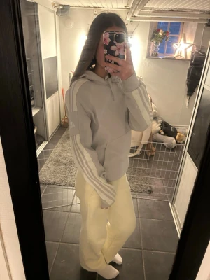 Ljusgrå Adidas hoodie med huva - Säljer en ljusgrå hoodie från Adidas med klassiska vita ränder längs ärmarna och dragsko i huvan. Hoodien har en avslappnad passform och är perfekt för en chill och sportig stil. Adidas-logga på ena ärmen.