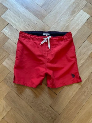 U.S Polo Assn badbyxor  - Snygga röda badbyxor från U.S Polo Assn med vit dragsko i midjan och broderad logga på vänster ben. Klassisk modell med elastisk midja och bakficka. Perfekta för stranden eller poolen.