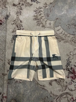 Beige Burberry shorts med rutigt mönster - Snygga beige shorts från Burberry med klassiskt rutigt mönster i grått och svart. De har elastisk midja med vit dragsko och sidofickor. Materialet känns lätt och passar perfekt till sommaren. Burberry-logga tryckt på vänster ben.