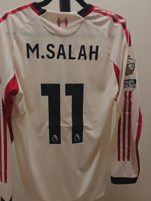 Liverpool Salah matchtröja långärmad - Vit långärmad Liverpool matchtröja med röda detaljer och svarta muddar. Tryck med M. Salah och nummer 11 på ryggen, Premier League-logga och 'No room for racism' på ärmen. Tillverkad i ventilerande funktionsmaterial, perfekt för fotbollsträning.