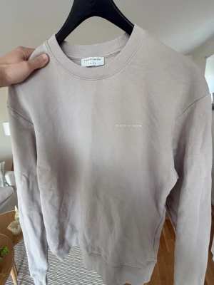 Beige sweatshirt från Tiger of Sweden - Stilren beige sweatshirt från Tiger of Sweden med rund halsringning och diskret logga på bröstet. Tröjan har en klassisk passform och ribbade muddar vid ärmslut och nederkant. Perfekt för en avslappnad och trendig look.