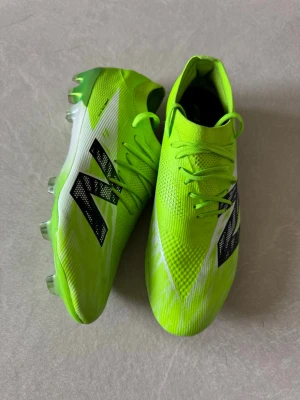 Neongröna New Balance fotbollsskor - Säljer ett par riktigt snygga neongröna fotbollsskor från New Balance med svart logga på sidan. Skorna har en strömlinjeformad design, snörning och ovandel i syntetmaterial för bra bollkontroll. Perfekta för dig som vill sticka ut på planen. Använda vid två tillfällen, kvittot finns vid önskemål och paketet följs med