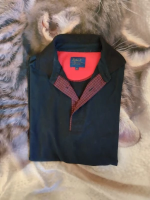 Svart henley-skjorta från Kyra K - Snygg svart henley-skjorta från Kyra K med röd och svart rutig detalj vid knäppningen och röd insida i kragen. Skjortan har klassisk krage och är perfekt för dig som gillar en stilren look med lite edge.